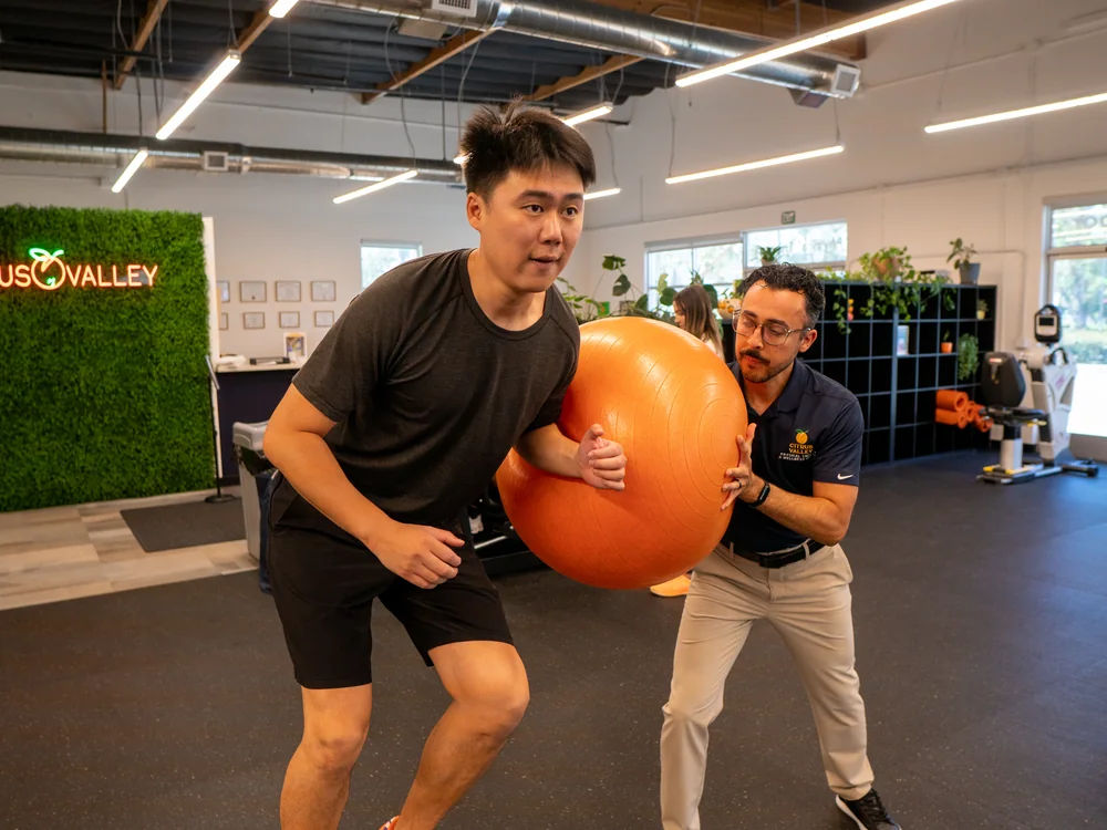 Patient squatting with an exercise ball while a therapist spots him