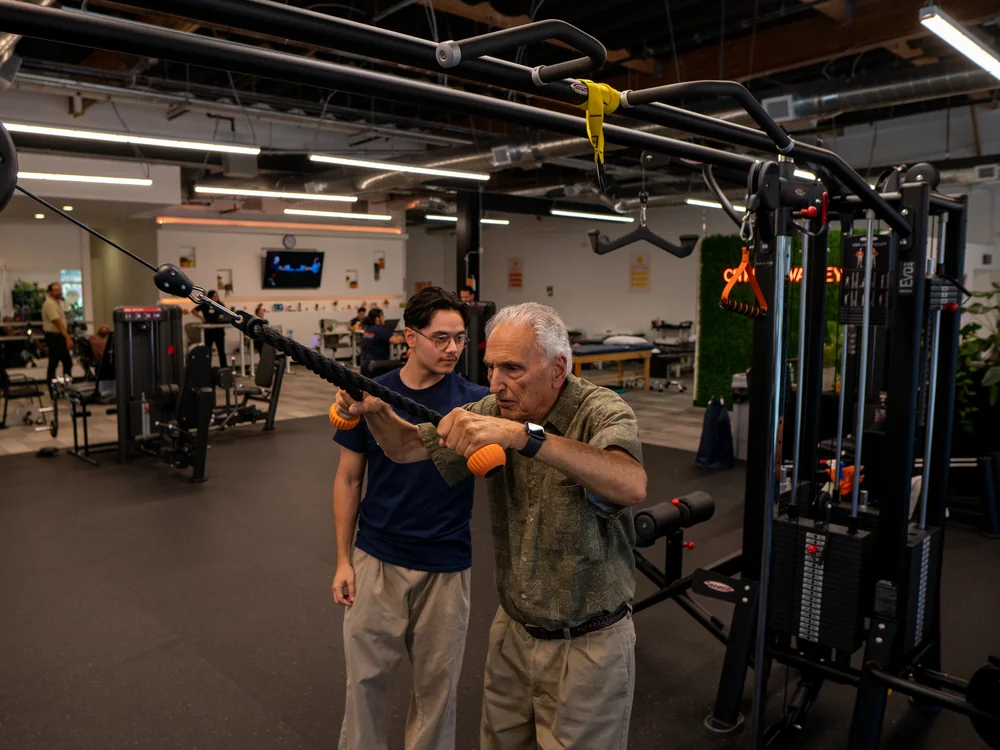 Older patient pulling cable handles on a resistance machine with a physical therapist beside him
